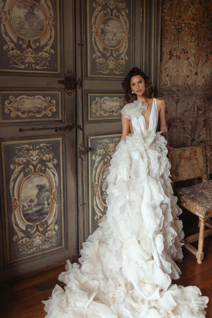 אישה בשמלה לבנה דרמטית עם מחשוף V עמוק עומדת ליד דלתות כפולות מעוטרות בסגנון וינטג' בחדר עם עיצוב עתיק וכיסא מעוטר. - Liel & Cavalli