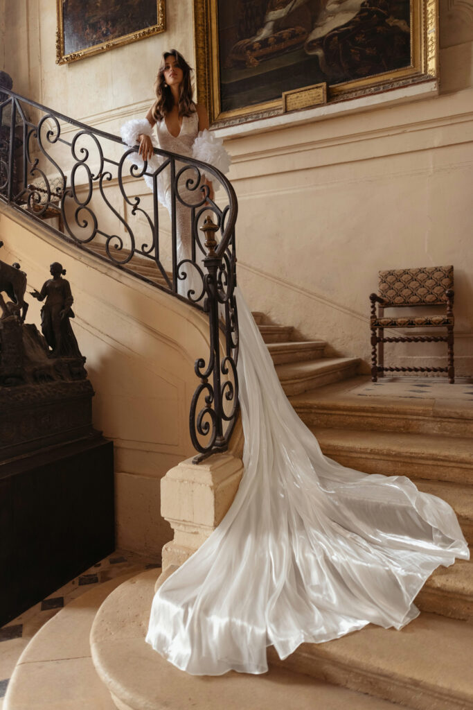 אישה בשמלה לבנה וזורמת עם שובל ארוך עומדת על גרם מדרגות מעוטר בפנים אלגנטיים, מוקפת באמנות קלאסית, פסלים ומעקות ברזל דקורטיביים. - Liel & Cavalli