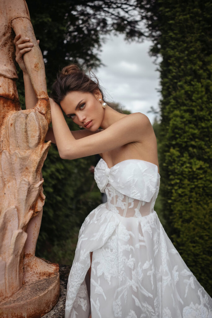 אישה בשמלת תחרה לבנה ללא כתפיות נשענת על פסל אבן בחוץ, מוקפת בצמחייה עבותה ועצים, ומביטה במצלמה במבט רך. - Liel & Cavalli