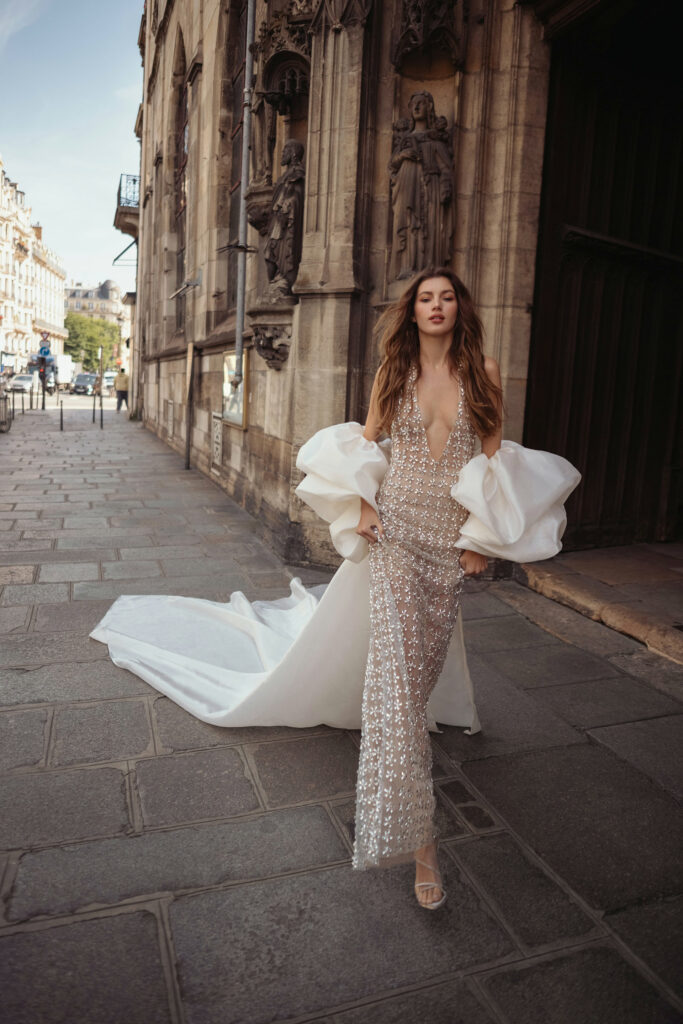 אישה בשמלה נוצצת עם מחשוף V עמוק, שרוולים לבנים דרמטיים ושובל ארוך, הולכת בביטחון ברחוב מרוצף אבן לצד בניין היסטורי מעוטר. - Liel & Cavalli