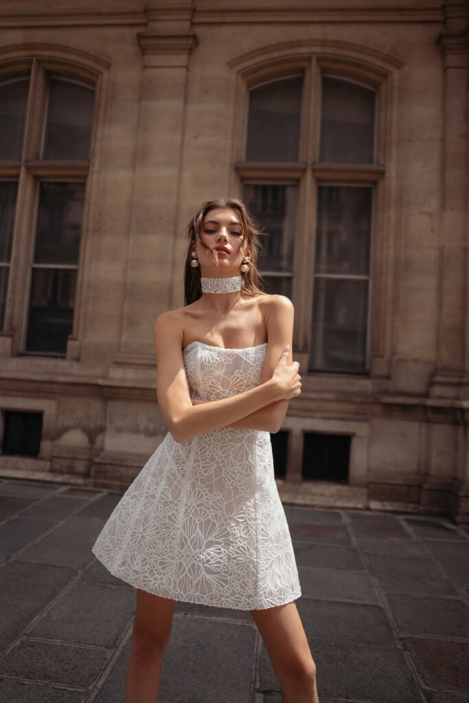 אישה בשמלת תחרה לבנה ללא כתפיות עם קולר תואם עומדת על מדרכה מאבן מול בניין עם חלונות גבוהים וקירות אבן בצבע בז'. זרועותיה שלובות באופן רופף והיא מביטה בביטחון למצלמה. - Liel & Cavalli