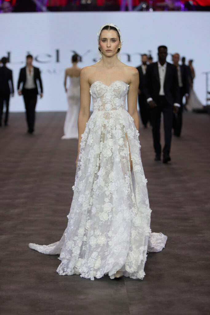 דוגמנית צועדת על מסלול תצוגה לבושה בשמלת כלה לבנה ללא כתפיות, מעוטרת בתחרה פרחונית, עם מחשוף לב ושובל עדין, כשברקע דוגמניות אחרות ומסך מואר. - Liel & Cavalli