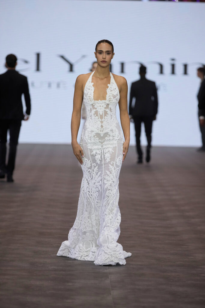 דוגמנית צועדת על המסלול לבושה בשמלת תחרה לבנה צמודה עם מחשוף עמוק ופרטים שקופים מורכבים. ברקע מוקרן טקסט מטושטש וצלליות של אנשים אחרים. - Liel & Cavalli