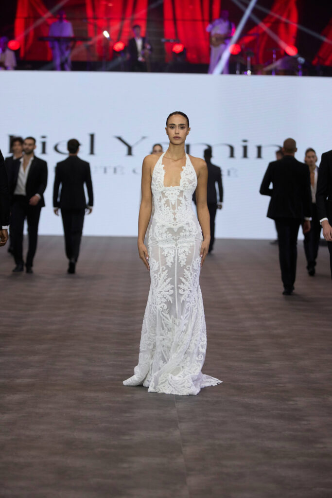 דוגמנית צועדת על המסלול לבושה בשמלת תחרה לבנה שקופה עם דוגמה מורכבת. גברים בחליפות כהות צועדים מאחוריה, עם מסך מואר גדול ותאורת במה אדומה ברקע. - Liel & Cavalli