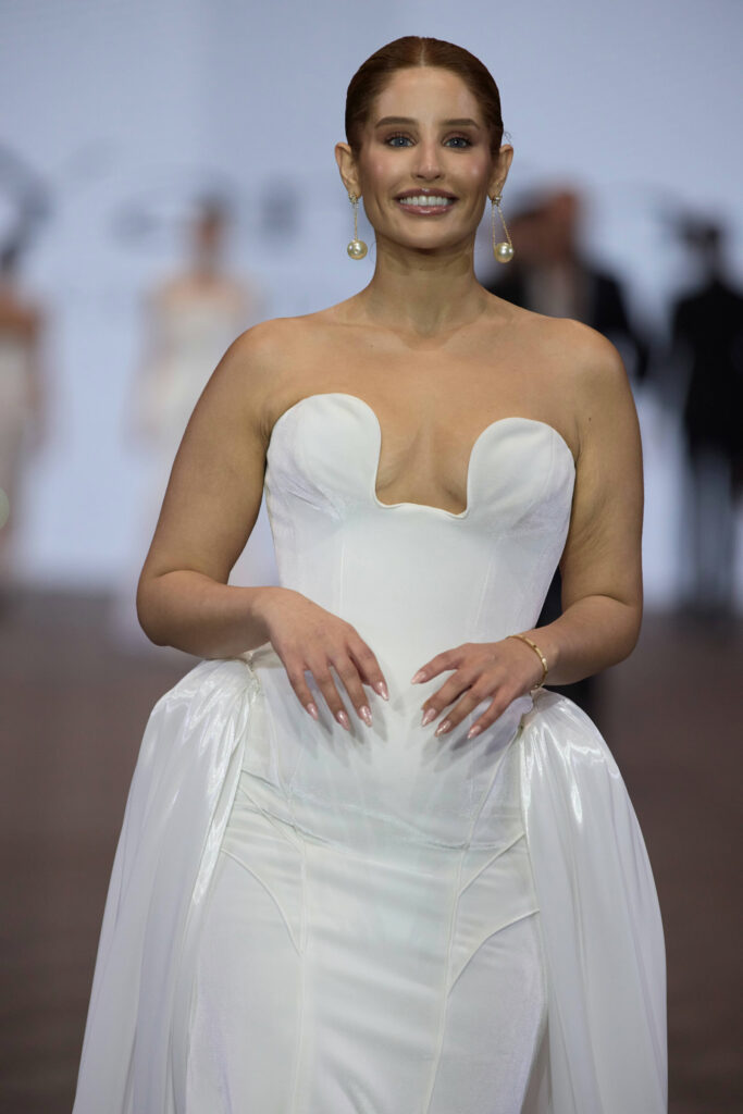 אישה בשמלה לבנה ללא כתפיות עם מחשוף לב מחייכת על מסלול תצוגת אופנה. שיערה מסורק לאחור, היא עונדת עגילים ארוכים ועומדת בביטחון עם ידיה שלובות. הרקע מטושטש עם צלליות. - Liel & Cavalli