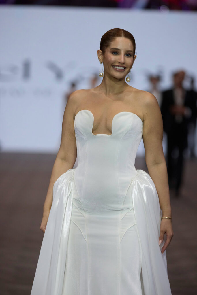 אישה עם שיער אדום מסורק לאחור לובשת שמלת ערב לבנה ללא כתפיות עם מחשוף מעוגל ייחודי ופאנלים צדדיים דרפתיים, מחייכת בזמן שהיא צועדת על המסלול. - Liel & Cavalli