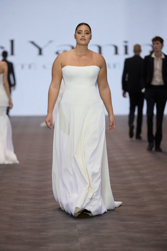 דוגמנית צועדת בביטחון על מסלול תצוגה לבושה בשמלת ערב לבנה ללא כתפיות. ברקע נראים מספר דוגמניות ואנשים נוספים, ומאחוריהם מסך מואר גדול. - Liel & Cavalli