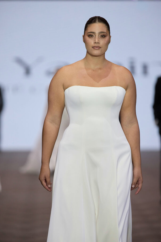 דוגמנית עם שיער כהה מסורק לאחור צועדת על המסלול לבושה בשמלת ערב לבנה ללא כתפיות באורך הרצפה. הרקע מטושטש, וניתן להבחין בטקסט וצלליות קלושות. - Liel & Cavalli