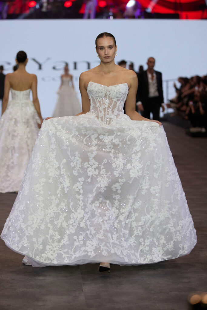 דוגמנית צועדת על המסלול בתצוגת אופנה בשמלת כלה לבנה ללא כתפיות בסגנון מחוך, עם חצאית נפוחה רקומה בתחרה. ברקע נראים דוגמניות אחרות וצופים. - Liel & Cavalli