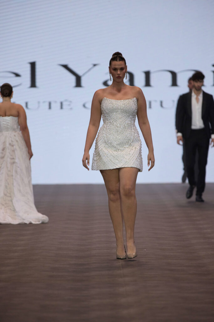 דוגמנית צועדת על מסלול תצוגה לבושה בשמלה קצרה, ללא כתפיות, לבנה ומעוטרת. ברקע נראות שתי דוגמניות נוספות, האחת בשמלה לבנה ארוכה והשנייה בחליפה שחורה. - Liel & Cavalli