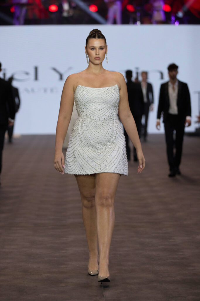 אישה לבושה בשמלה קצרה, ללא כתפיות, עם חרוזים לבנים ועקבים שקופים, צועדת בביטחון על מסלול תצוגה, כשברקע מספר אנשים ומסך מואר. - Liel & Cavalli