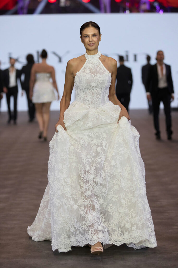 דוגמנית צועדת על מסלול תצוגה לבושה בשמלת תחרה לבנה ללא שרוולים, עם צווארון גבוה וחצאית מלאה. ברקע נראים דוגמניות אחרות ומסך מטושטש עם טקסט. - Liel & Cavalli