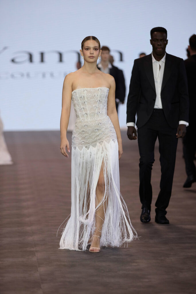 דוגמנית צועדת על המסלול בשמלה לבנה ללא כתפיות, מעוטרת בחרוזים, עם פרנזים וחתך גבוה. היא נועלת נעלי עקב בצבע גוף. מאחוריה צועד דוגמן בחליפה שחורה וחולצה לבנה על מסלול תצוגת האופנה. - Liel & Cavalli