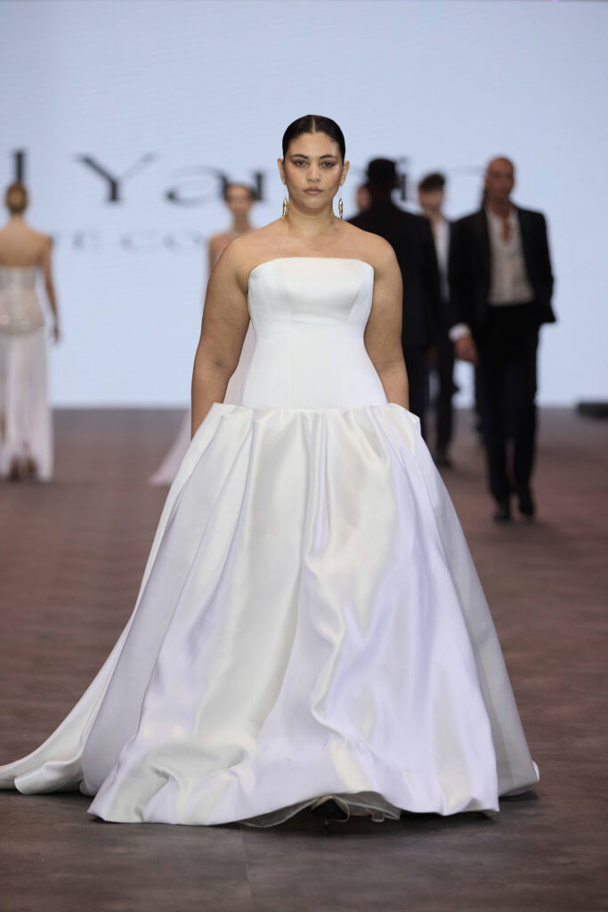 דוגמנית צועדת על המסלול בשמלת כלה לבנה ללא כתפיות, עם חצאית מלאה ועיטורים מינימליים. ברקע, על מסלול תצוגת האופנה, נראות דוגמניות אחרות לבושות בבגדים אלגנטיים. - Liel & Cavalli