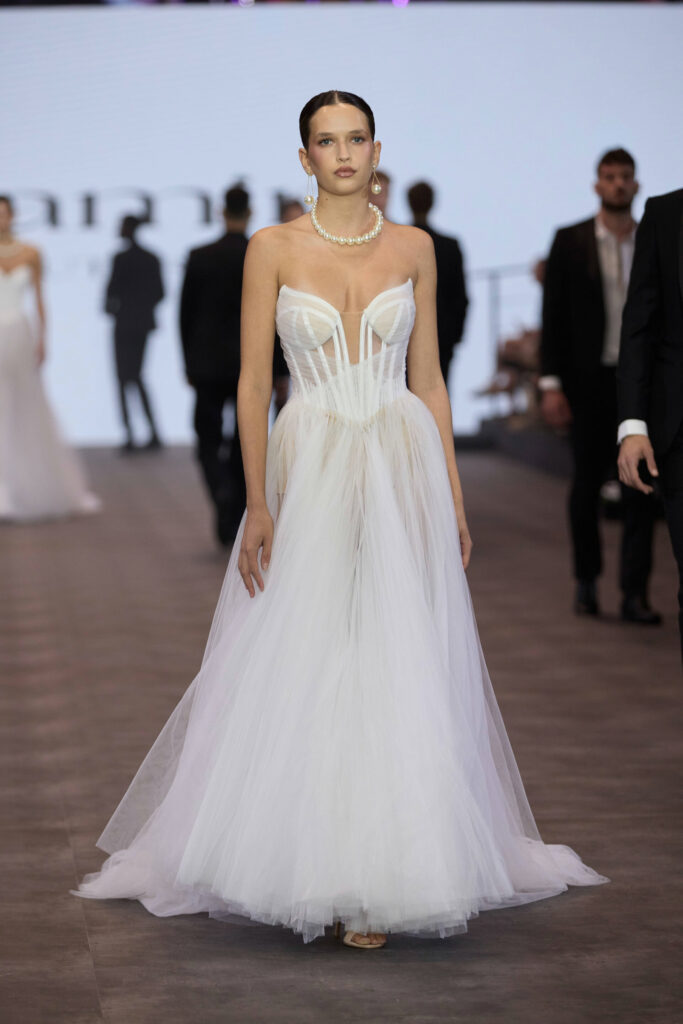 דוגמנית צועדת על מסלול תצוגה לבושה בשמלת טול לבנה ללא כתפיות, עם מחוך מחויט וחצאית מלאה. היא משלימה את המראה עם שרשרת פנינים ועגילים. ברקע נראים אנשים לבושים בבגדים רשמיים. - Liel & Cavalli