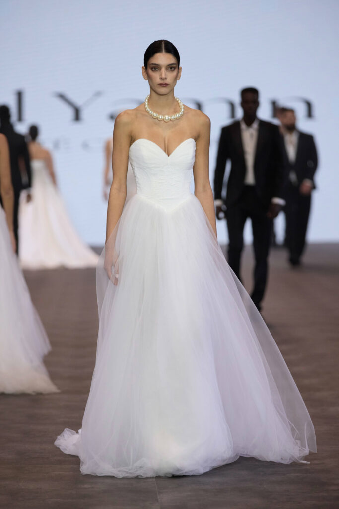 דוגמנית צועדת על המסלול לבושה בשמלת כלה לבנה ללא כתפיות עם מחשוף לב וחצאית טול, משלימה את המראה עם שרשרת פנינים גדולה. ברקע נראות דוגמניות נוספות לבושות בבגדים רשמיים. - Liel & Cavalli