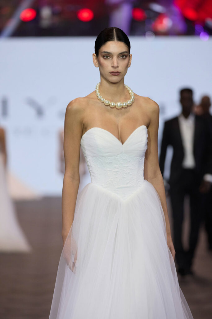 דוגמנית עם שיער משוח לאחור לובשת שמלת כלה לבנה ללא כתפיות עם מחשוף לב ושרשרת פנינים, והולכת על מסלול תצוגה. הרקע מטושטש וניתן לראות דוגמניות אחרות ואורות. - Liel & Cavalli
