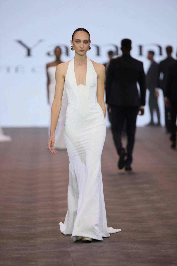 דוגמנית לבושה בשמלה לבנה ואלגנטית עם מחשוף סטרפלס צועדת על מסלול תצוגה. ברקע נראים דוגמניות אחרות ומסך מטושטש עם טקסט. נראה כי התמונה צולמה בתצוגת אופנה. - Liel & Cavalli