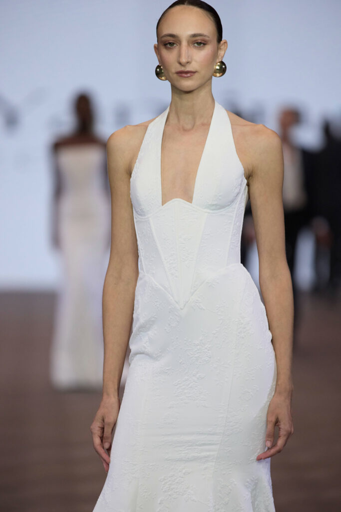 דוגמנית עם שיער משוח לאחור ועגילי זהב גדולים צועדת על המסלול בשמלת ערב לבנה צמודה עם מחשוף סטרפלס ועם דוגמה מרקמית עדינה. הרקע מטושטש, ומראים בו דמויות אחרות. - Liel & Cavalli