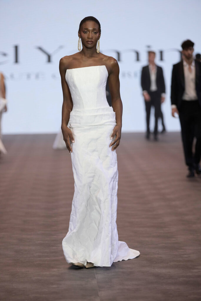 דוגמנית צועדת בביטחון על המסלול לבושה בשמלה לבנה ללא כתפיות, באורך הרצפה, עם מרקם מקומט עדין. ברקע נראות דמויות מטושטשות ומסך בהיר המציג טקסט. - Liel & Cavalli