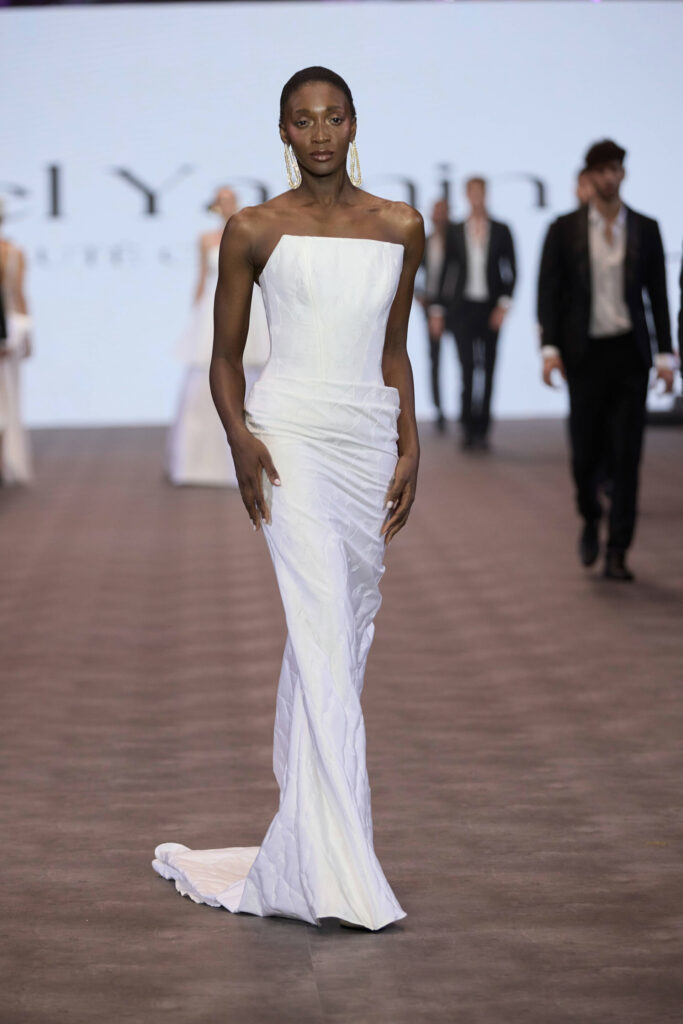 דוגמנית צועדת על המסלול לבושה בשמלת ערב לבנה ללא כתפיות, באורך הרצפה, עם שובל קל, בתצוגת אופנה עם דמויות מטושטשות ברקע. - Liel & Cavalli