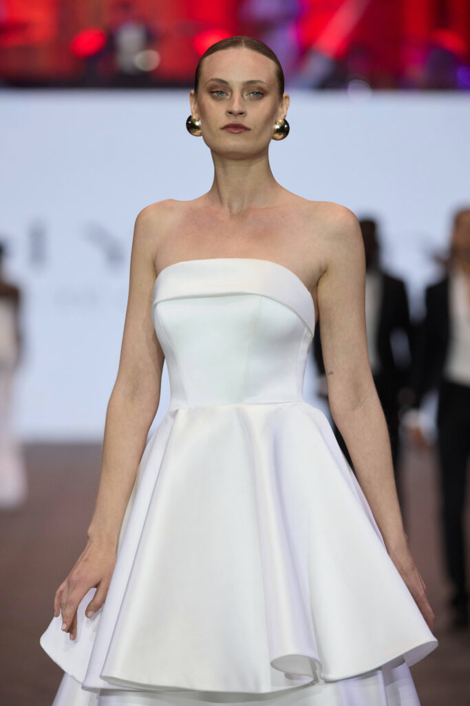 דוגמנית לבושה בשמלה לבנה, ללא כתפיות, עם עגילי חישוק זהב, צועדת על המסלול. הרקע מטושטש, עם אנשים ואורות צבעוניים. - Liel & Cavalli