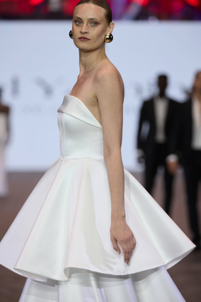 דוגמנית בשמלה לבנה מחויטת ללא כתפיות עם חצאית מתרחבת צועדת על מסלול תצוגת אופנה. היא עונדת עגילי זהב גדולים ושיערה אסוף לאחור. הרקע מטושטש, ומראים בו אנשים בלבוש רשמי. - Liel & Cavalli