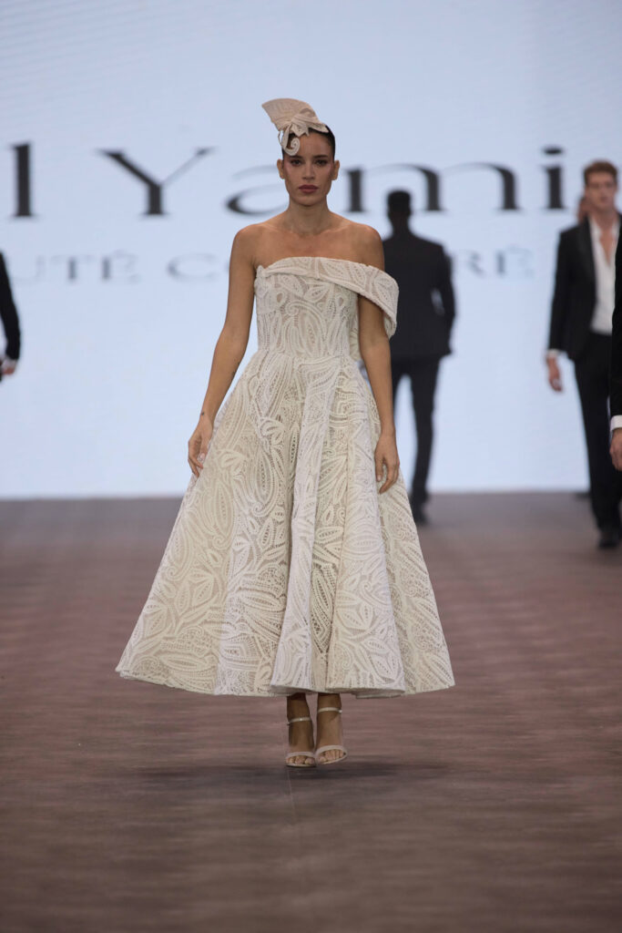 דוגמנית צועדת על מסלול תצוגה לבושה בשמלת מידי בצבע שמנת עם הדפס, עם כתפיים חשופות, וכובע תואם. ברקע מסך מואר עם טקסט מטושטש ואנשים אחרים בחליפות. - Liel & Cavalli