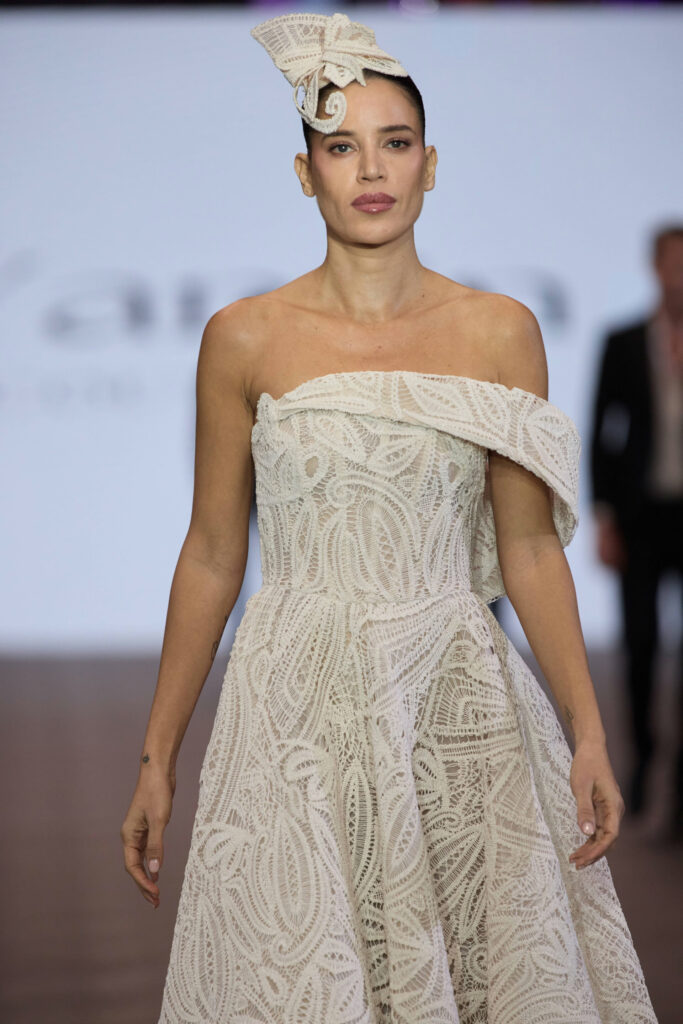 דוגמנית צועדת על מסלול תצוגה לבושה בשמלה לבנה עם כתפיים חשופות ועיטורי תחרה, עם כיסוי ראש תואם, על רקע מטושטש. - Liel & Cavalli