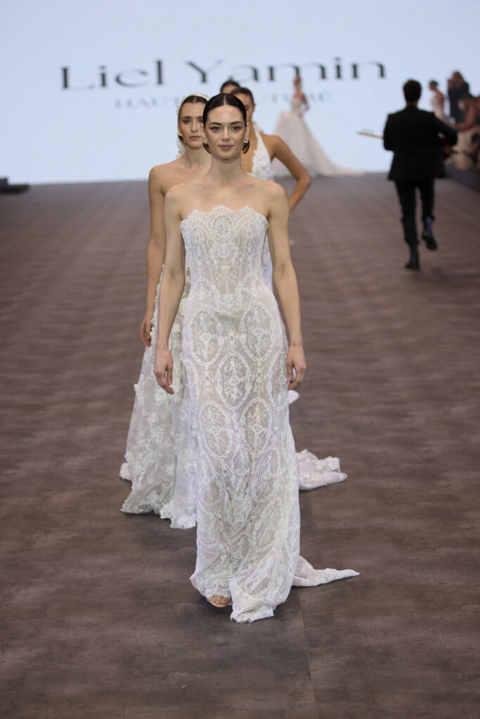 דוגמנית לבושה בשמלת תחרה לבנה ללא כתפיות, בעלת דוגמה מורכבת, צועדת על מסלול תצוגת אופנה, ואחריה דוגמניות אחרות בשמלות דומות. ברקע מופיע שם המעצב. - Liel & Cavalli