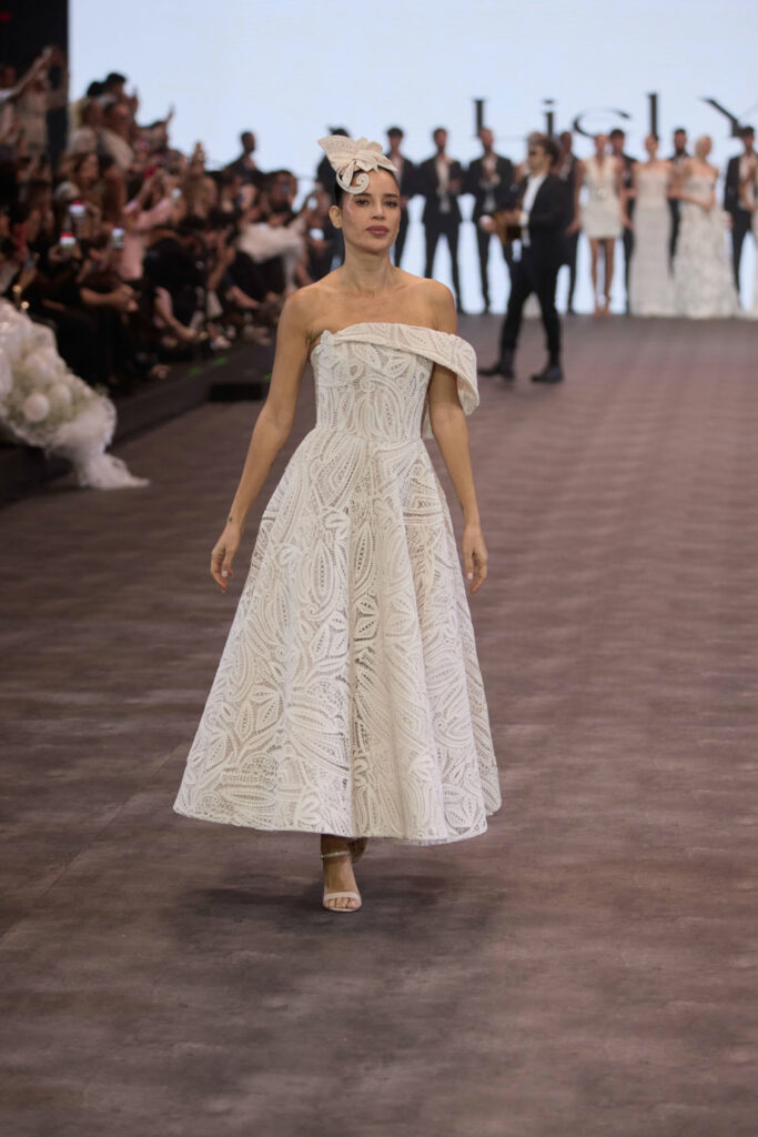 דוגמנית צועדת על מסלול תצוגה לבושה בשמלה לבנה באורך הברך עם כתפיים חשופות, בעלת דוגמה מרקמת וכובע לבן. הקהל צופה, ודוגמניות אחרות עומדות ברקע. - Liel & Cavalli
