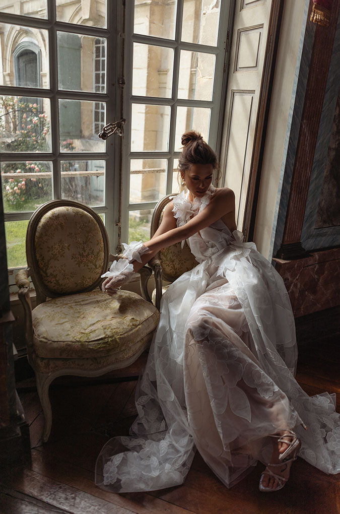 אישה בשמלה לבנה מתנפנפת יושבת על כיסא מעוטר ליד חלון גדול, כשקרני השמש חודרות פנימה וגן נראה מבחוץ. היא מביטה מטה, מסדרת את הכפפה שלה, בחדר אלגנטי ומואר באור רך. - Liel & Cavalli