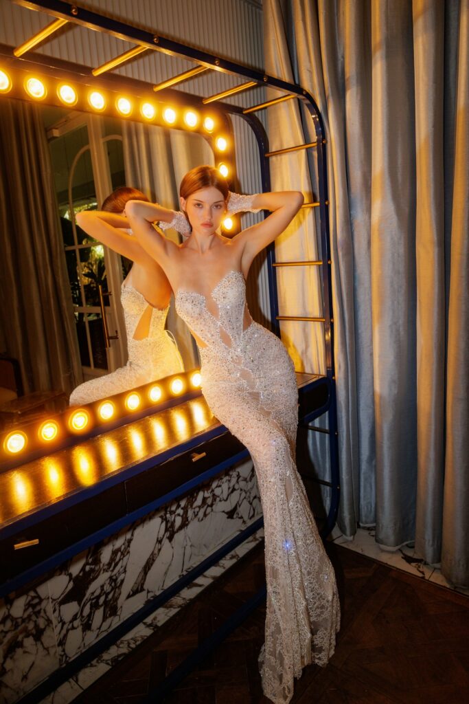 אישה בשמלה נוצצת וצמודה לדמותה מצטלמת מול מראה גדולה ומוארת עם משטח שיש ווילונות כחולים ברקע. היא מניחה את ידיה על ראשה ומביטה בביטחון למצלמה. - Liel & Cavalli