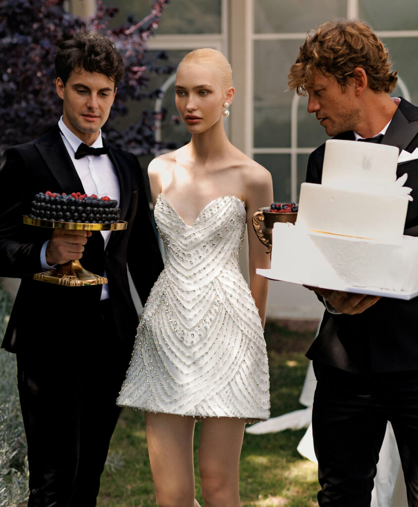 שלושה אנשים בחתונה תחת כיפת השמיים: אישה בשמלה לבנה קצרה ומחורזת עומדת בין שני גברים בחליפות טוקסידו המחזיקים קינוחים — האחד עם עוגת פירות יער והשני עם עוגה לבנה רב-קומתית. - Liel & Cavalli