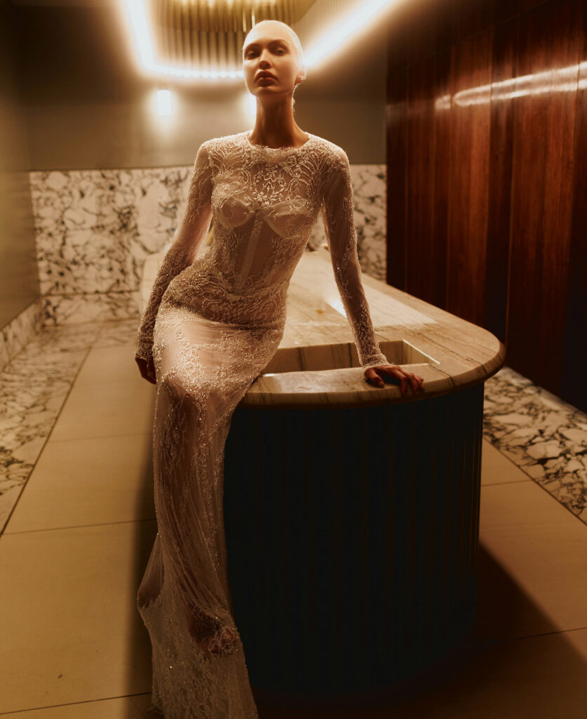 אישה בשמלה ארוכת שרוולים שקופה ומרוקמת בחרוזים מורכבים נשענת על דלפק שיש בחדר מודרני ומואר באור עמום, עם קירות שיש ולוחות עץ. - Liel & Cavalli