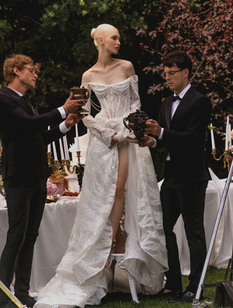 אישה בשמלת תחרה לבנה מהודרת עומדת בחוץ בין שני גברים בחליפות המחזיקים גביעים מעוטרים. מאחוריה, שולחן מעוטר במנורות, פרחים ונרות, המוצב בגן שופע. - Liel & Cavalli