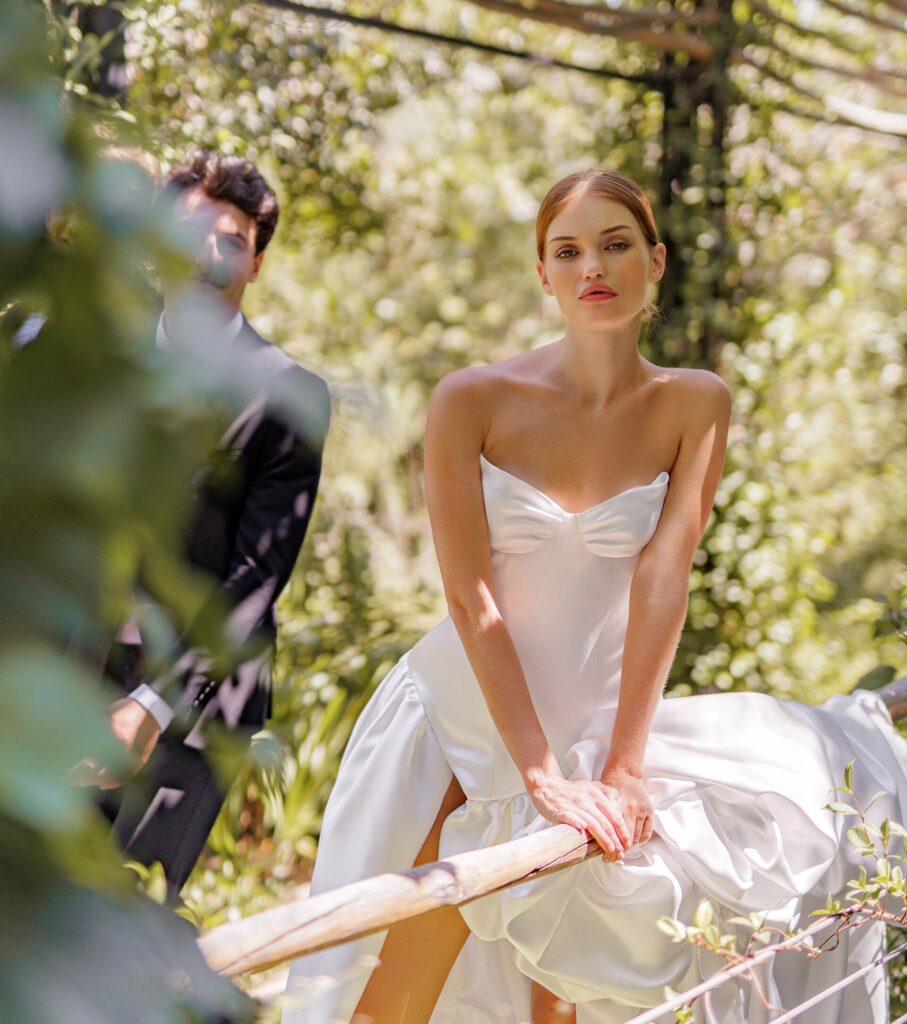 אישה בשמלת כלה לבנה ללא כתפיות נשענת על מעקה עץ בחוץ, מוקפת בצמחייה. גבר בחליפה שחורה עומד ברקע, מוסתר חלקית על ידי עלים. - Liel & Cavalli