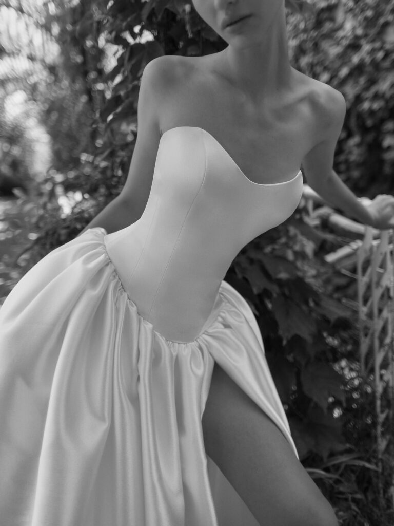 אישה בשמלת סאטן ללא כתפיות עם חתך עד הירך מצטלמת בחוץ ליד עלווה. התמונה בשחור-לבן, מה שמדגיש את המרקם החלק והעיצוב האלגנטי של השמלה. - Liel & Cavalli