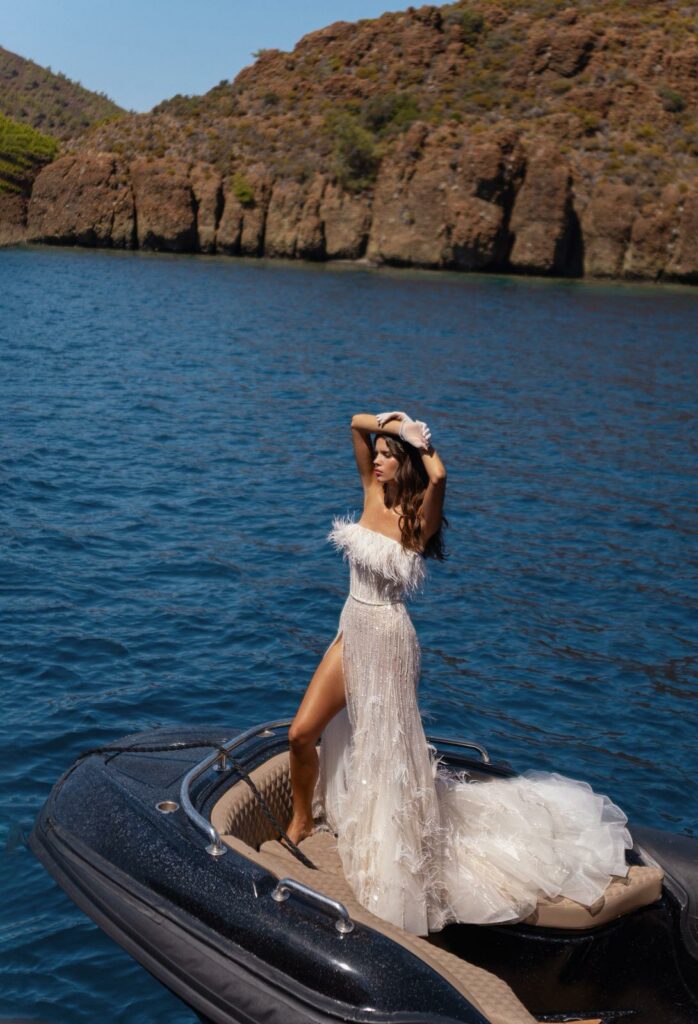 אישה בשמלת נוצות לבנה ללא כתפיות עומדת על סירה שחורה קטנה במים כחולים, ומצטלמת עם ידיה על ראשה. ברקע, תחת שמיים בהירים, נראים צוקים סלעיים וצמחייה ירוקה. - Liel & Cavalli