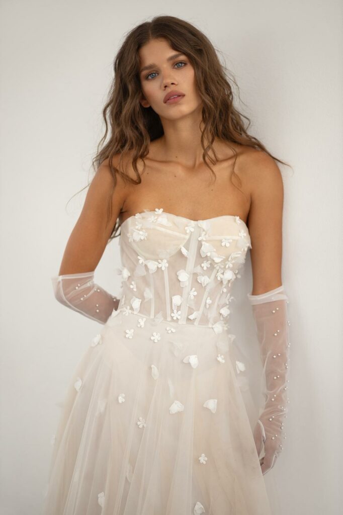 אישה עם שיער חום ארוך וגלי לובשת שמלה לבנה ללא כתפיות מעוטרת באפליקציות פרחוניות קטנות וכפפות שקופות עם פרטים פנינים, עומדת על רקע לבן חלק. - Liel & Cavalli