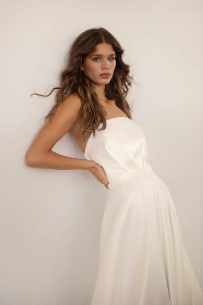 אישה עם שיער חום ארוך וגלי לובשת שמלת סאטן לבנה ללא כתפיות ומצטלמת על רקע קיר בהיר וחלק, מביטה במצלמה במבט רגוע ורציני. - Liel & Cavalli