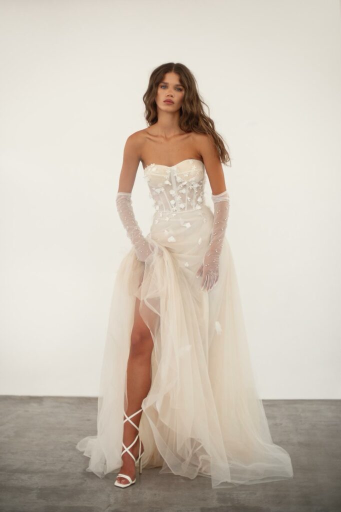 אישה בשמלת סטרפלס שקופה בצבע שנהב עם מחוך מעוטר באפליקציות פרחוניות לבנות עומדת על רצפה אפורה. היא לובשת כפפות טול ארוכות, נעלי עקב לבנות עם שרוכים, ויש לה שיער ארוך וגלי. - Liel & Cavalli