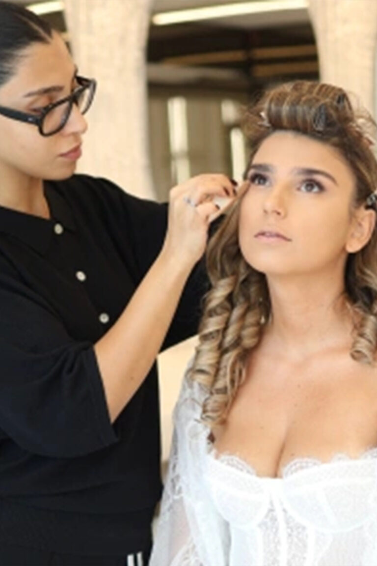 מאפרת המרכיבה משקפיים מאפרת אישה עם שיער מתולתל, הלובשת חלוק לבן ומביטה כלפי מעלה בחדר מואר היטב. - Liel & Cavalli