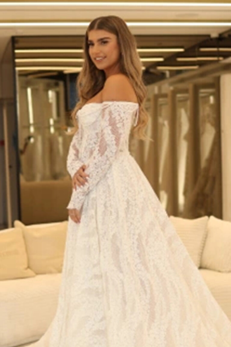 אישה עם שיער ארוך וגלי לובשת שמלת כלה לבנה מתחרה עם כתפיים חשופות, עומדת בתוך הבית, מחייכת קלות, עם ספות בצבע שמנת ברקע. - Liel & Cavalli