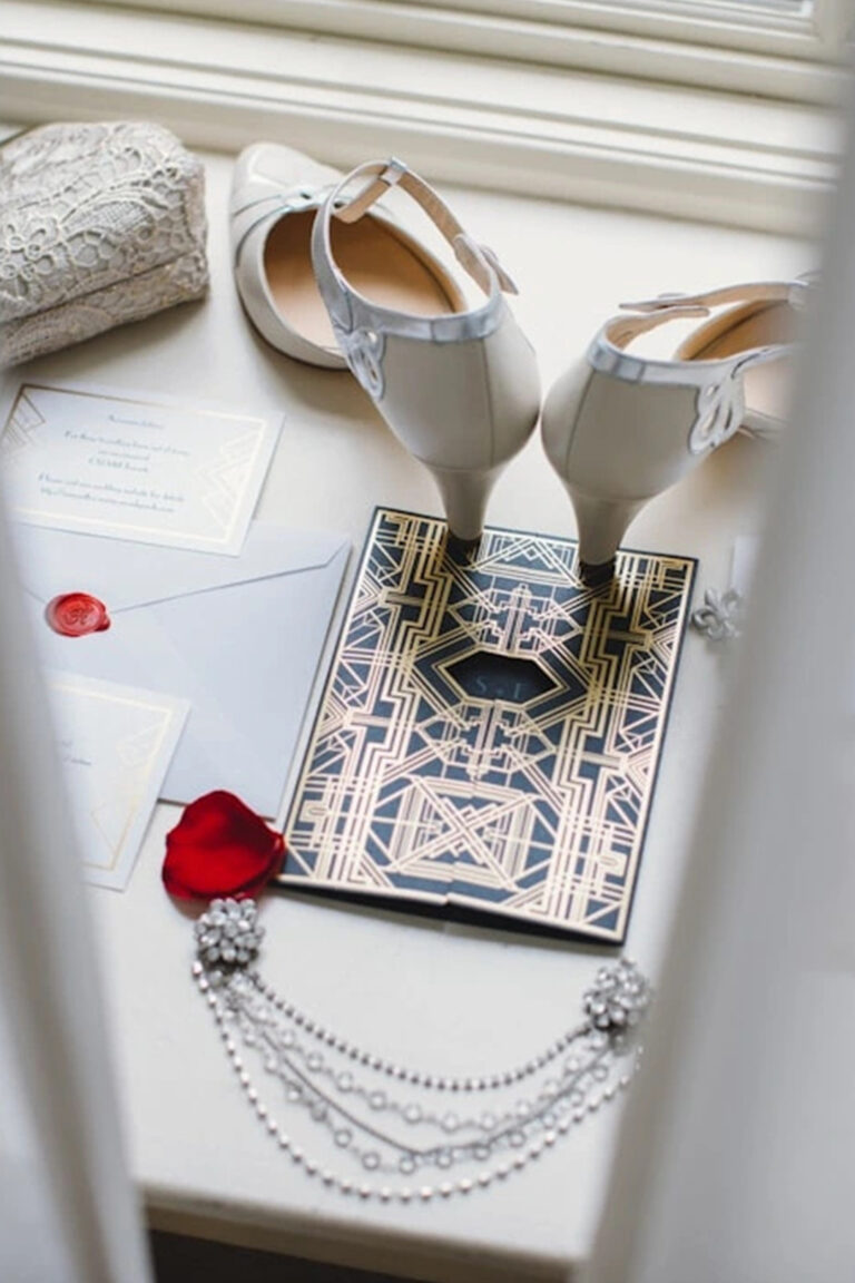 זוג נעליים לבנות עם עקבים גבוהים, תיק תחרה, הזמנה בסגנון ארט דקו, שרשרת, עלי כותרת של ורד אדום ומעטפה עם חותמת אדומה מסודרים על משטח לבן ליד חלון. - Liel & Cavalli